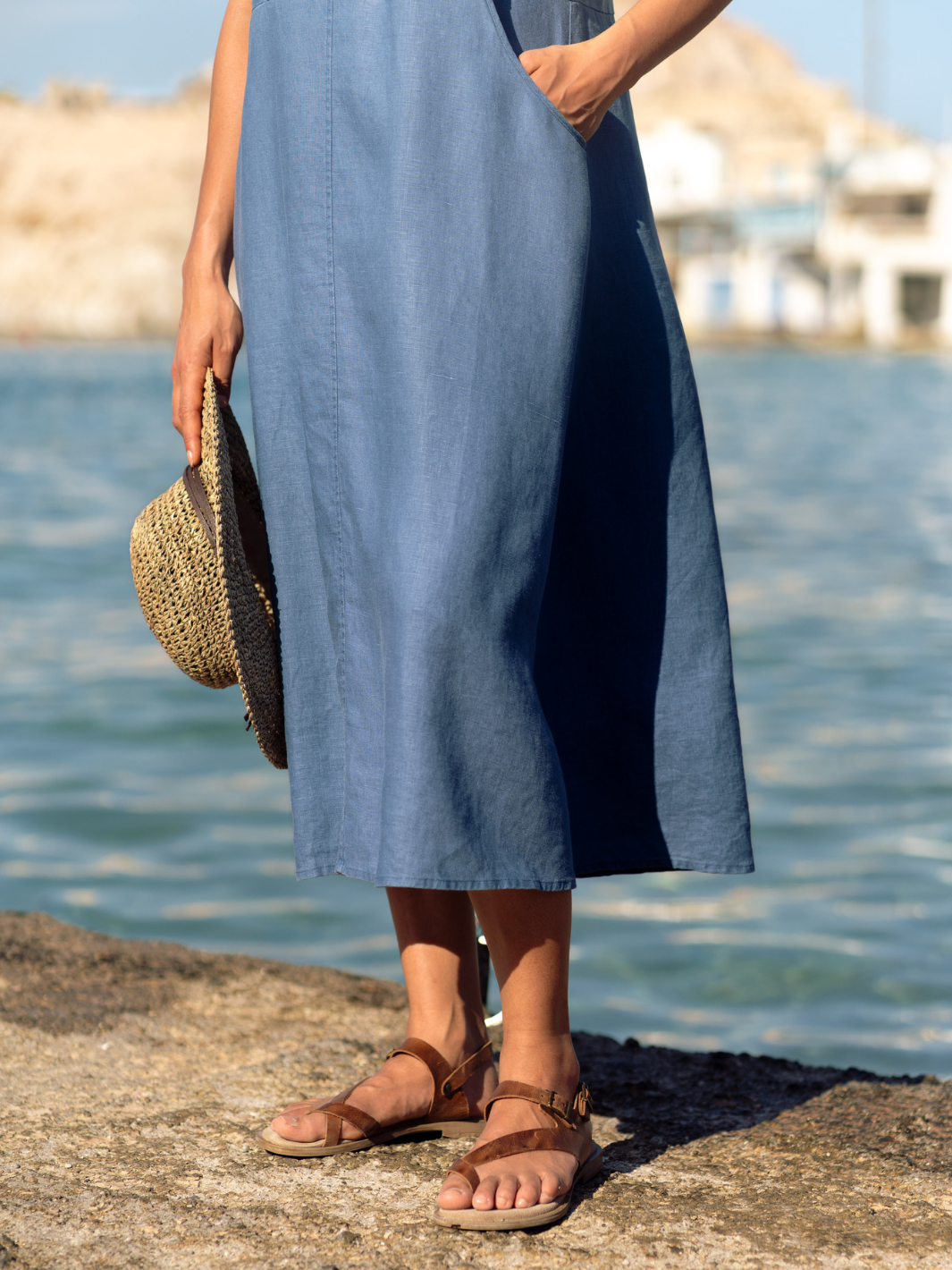 Woman in a blue linen dress standing by the sea holding a woven hat