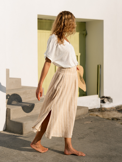 Woman in a white linen top and beige cotton skirt standing by the sea holding a woven hat