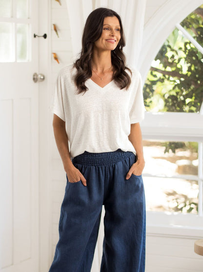 Woman wearing a white top and blue pants standing indoors with a window in the background.