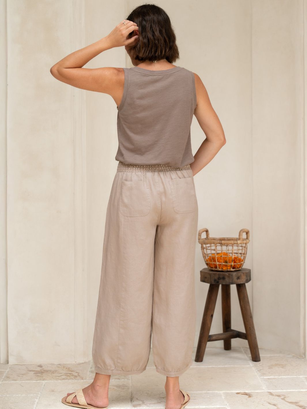 Woman wearing a sleeveless top and beige pants standing in a room with a wooden stool and basket in the background.