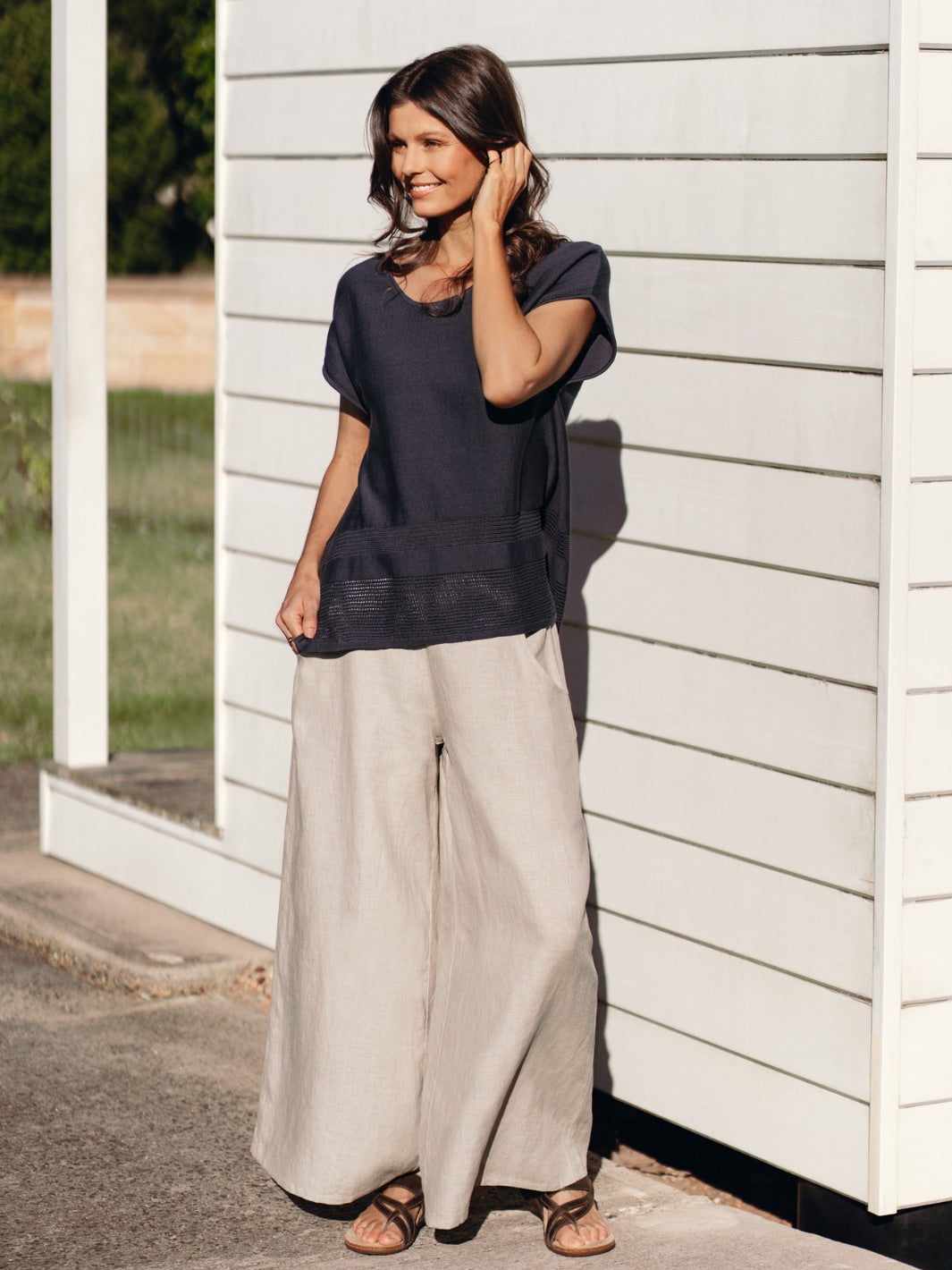 Woman wearing a dark top and light pants standing against a white wall.
