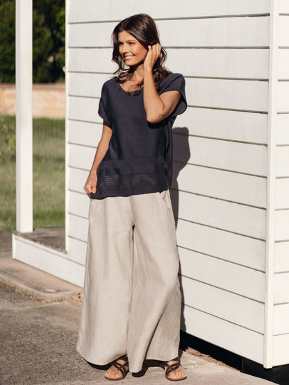 Woman wearing a dark top and light pants standing against a white wall.