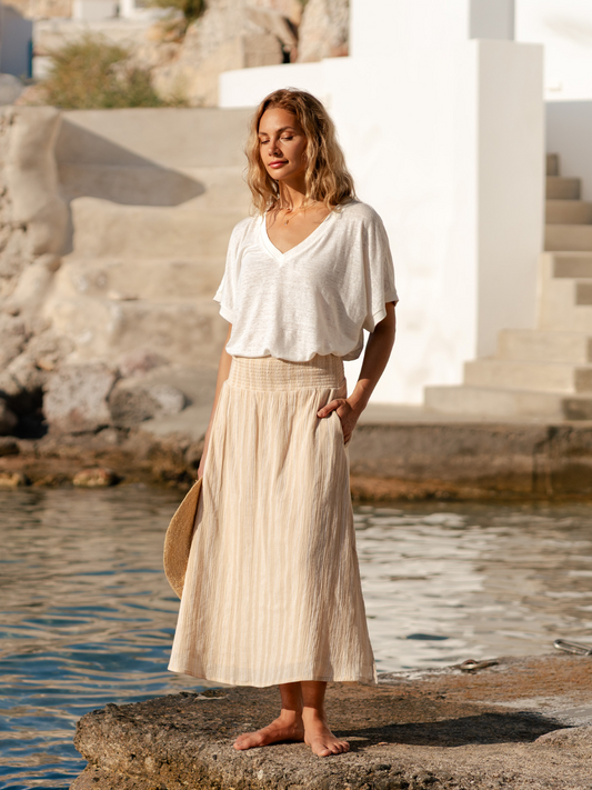 Woman in a white linen top and beige cotton skirt standing by the sea holding a woven hat