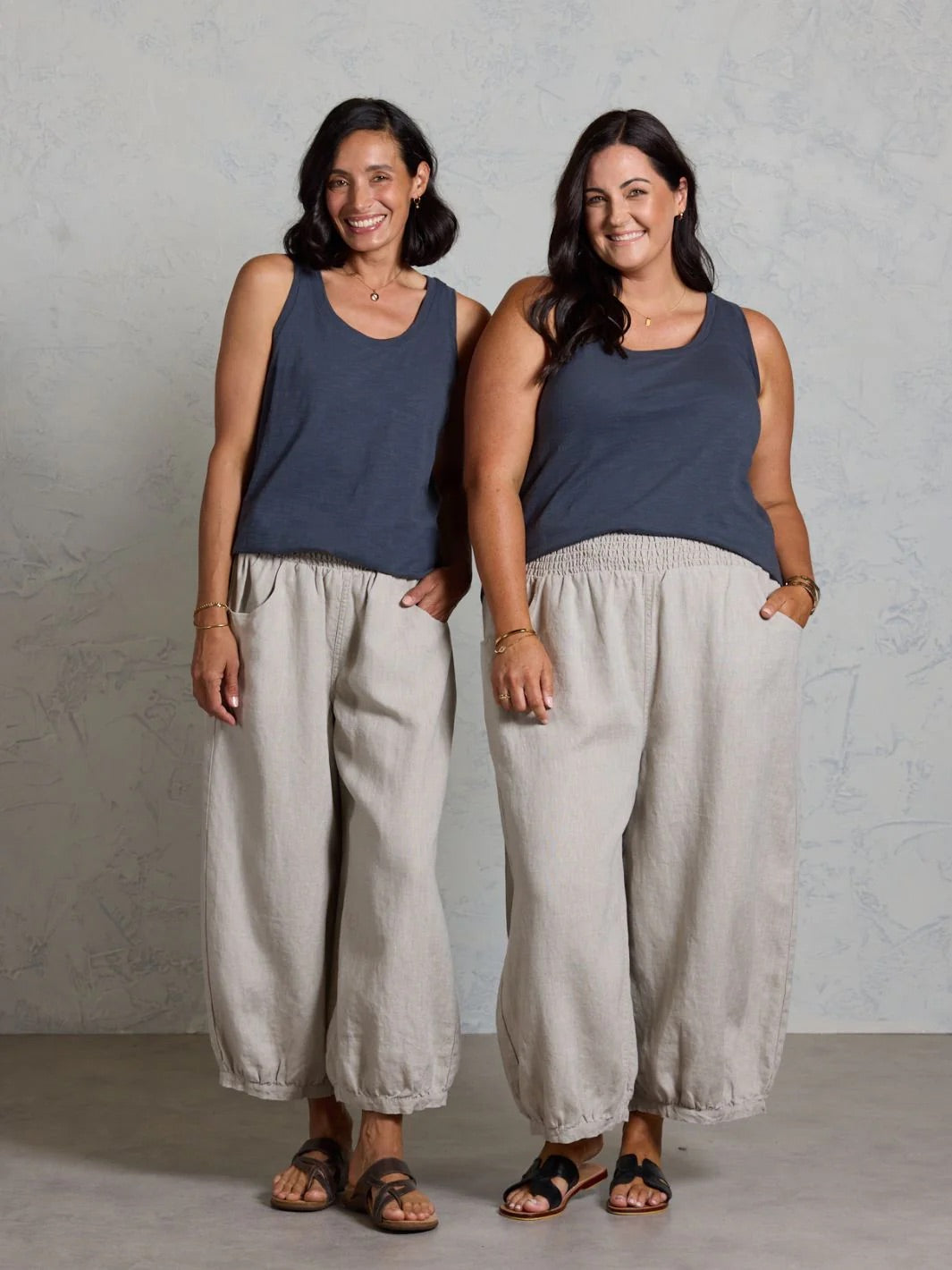 Two women wearing matching tank tops and beige pants against a plain background